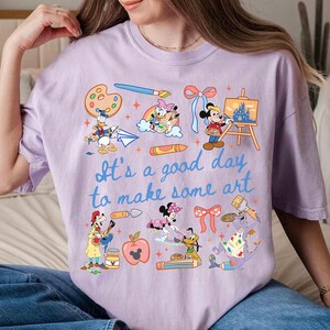 May include: A light purple t-shirt featuring various Disney characters and art supplies. The shirt includes the text "It's a good day to make some art" in blue script. Donald Duck, Mickey Mouse, and other characters are depicted.