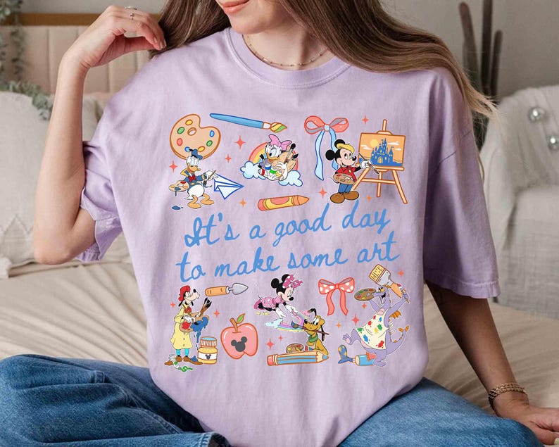 May include: A light purple t-shirt featuring various Disney characters and art supplies. The shirt includes the text "It's a good day to make some art" in blue script. Donald Duck, Mickey Mouse, and other characters are depicted.