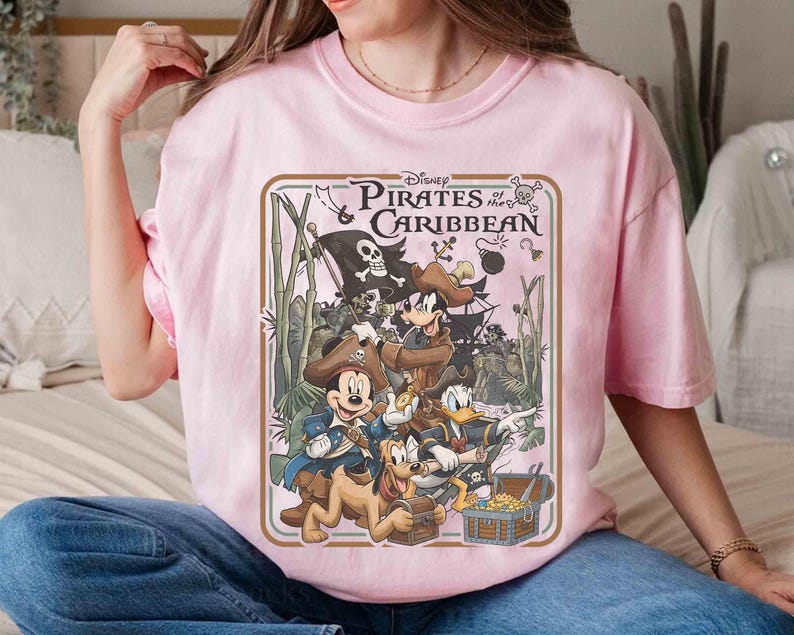 May include: A light pink t-shirt featuring a Disney Pirates of the Caribbean graphic. The graphic includes Mickey Mouse, Donald Duck, and Goofy dressed as pirates, along with a treasure chest and pirate ship. The text "Disney Pirates of the Caribbean" is displayed at the top.