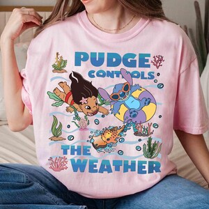 May include: Pink t-shirt featuring a graphic of two cartoon characters, one wearing sunglasses in a yellow and purple inner tube, and another swimming. The text "Pudge Controls The Weather" is printed in blue.