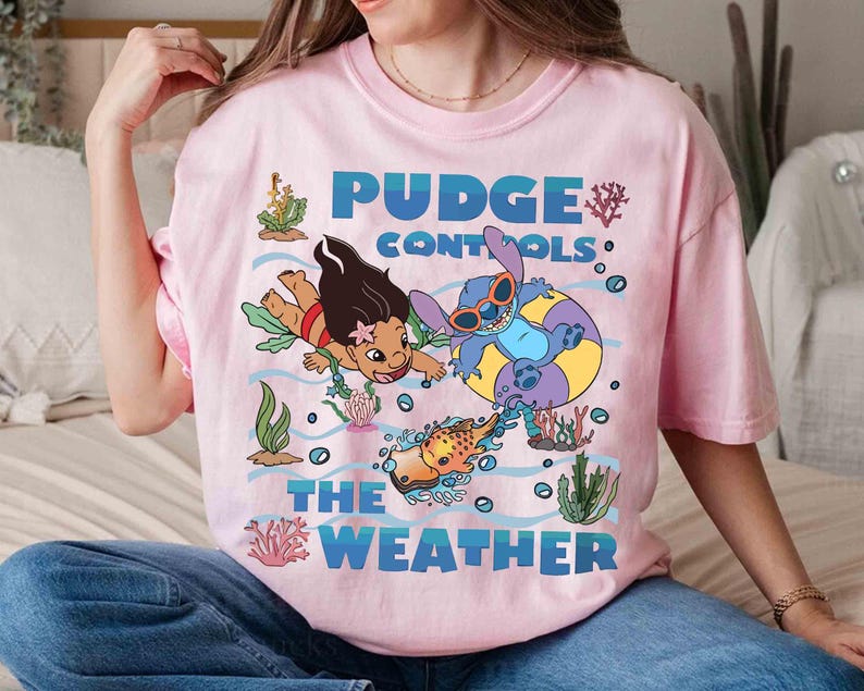 May include: Pink t-shirt featuring a graphic of two cartoon characters, one wearing sunglasses in a yellow and purple inner tube, and another swimming. The text "Pudge Controls The Weather" is printed in blue.