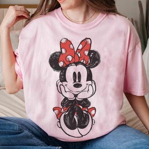 May include: A pale pink t-shirt with a black and white Minnie Mouse illustration. Minnie Mouse is wearing a red bow with white polka dots. The t-shirt has a loose fit.