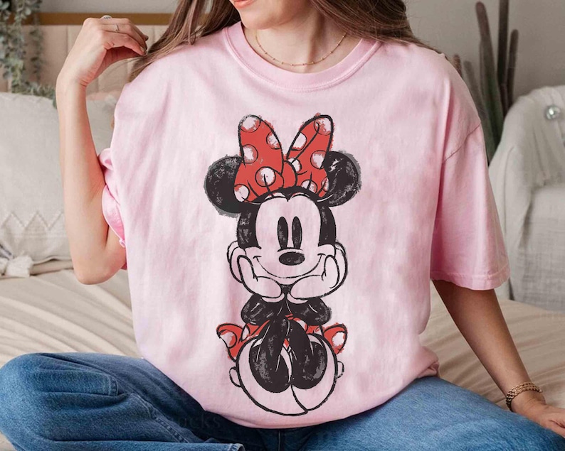 May include: A pale pink t-shirt with a black and white Minnie Mouse illustration. Minnie Mouse is wearing a red bow with white polka dots. The t-shirt has a loose fit.