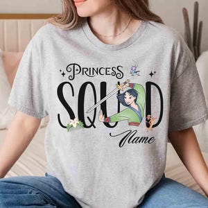 May include: A light gray t-shirt featuring the text "Princess Squad" in black script with a cartoon illustration of a woman holding a sword. The shirt also includes the word "Name" in script.
