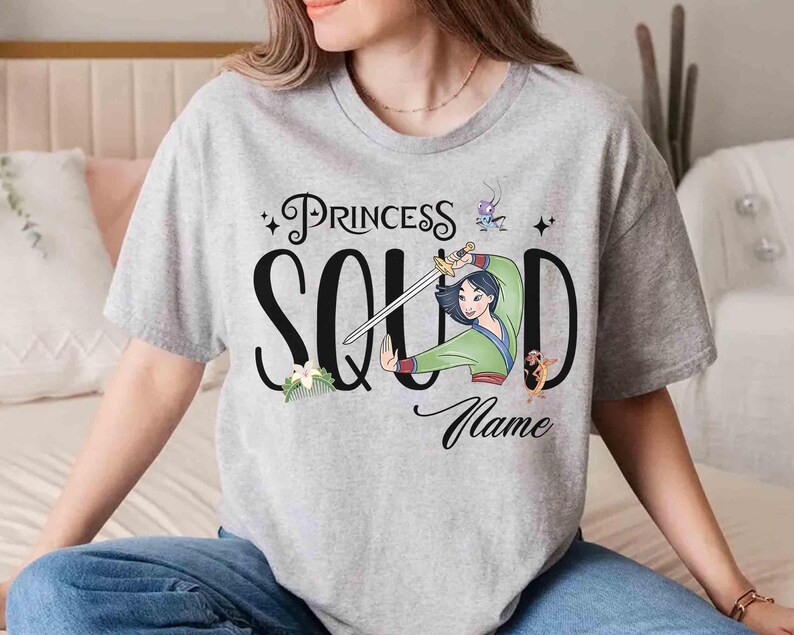 May include: A light gray t-shirt featuring the text "Princess Squad" in black script with a cartoon illustration of a woman holding a sword. The shirt also includes the word "Name" in script.