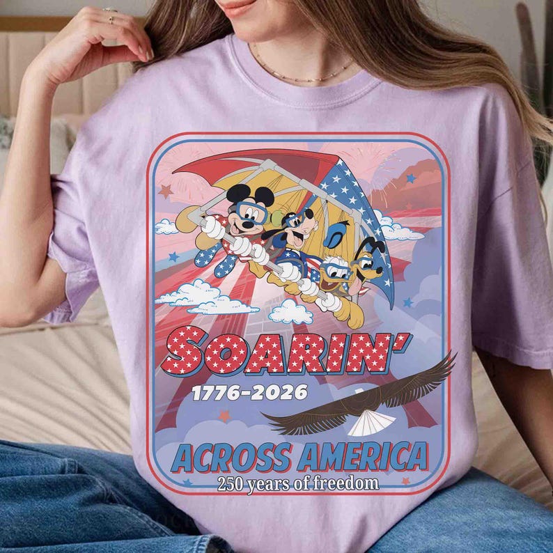 May include: A light purple t-shirt featuring a graphic of Mickey Mouse, Goofy, and Donald Duck hang gliding. The graphic includes the text "SOARIN' 1776-2026 ACROSS AMERICA 250 years of freedom" with an eagle illustration.