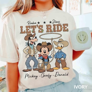 May include: Ivory-colored t-shirt featuring a rodeo theme with Mickey Mouse, Goofy, and Donald Duck dressed as cowboys. The shirt has the text "Rodeo Stars LET'S RIDE" above the characters and the names "Mickey, Goofy, Donald" below. A white mug with a smiley face is also visible.