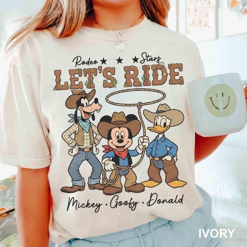 May include: Ivory-colored t-shirt featuring a rodeo theme with Mickey Mouse, Goofy, and Donald Duck dressed as cowboys. The shirt has the text "Rodeo Stars LET'S RIDE" above the characters and the names "Mickey, Goofy, Donald" below. A white mug with a smiley face is also visible.