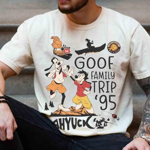 Pode incluir: Camiseta creme com um gr&aacute;fico de Pateta e Max da s&eacute;rie de televis&atilde;o Goof Troop da Disney. O design inclui o texto "GOOF FAMILY TRIP '95" e ilustra&ccedil;&otilde;es de viagem. A camiseta tem mangas curtas.