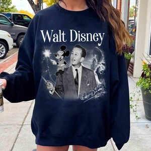 Puede incluir: Una sudadera de cuello redondo en azul marino oscuro con un estampado gr&aacute;fico de Walt Disney en traje y corbata, con Mickey Mouse sobre su hombro. El texto Walt Disney est&aacute; impreso en blanco sobre la imagen, con una firma en la parte inferior.