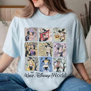 May include: Pale blue t-shirt with a grid of nine Disney princesses dressed as witches for Halloween. The text "Walt Disney World" is printed at the bottom. Each princess is depicted with Halloween-related imagery, such as pumpkins and cobwebs.