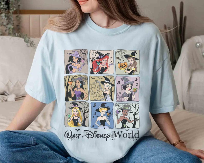 May include: Pale blue t-shirt with a grid of nine Disney princesses dressed as witches for Halloween. The text "Walt Disney World" is printed at the bottom. Each princess is depicted with Halloween-related imagery, such as pumpkins and cobwebs.