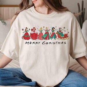 May include: Cream-colored t-shirt featuring a festive design with several princesses in holiday attire. The princesses are depicted in colorful dresses, with the text "MERRY CHRISTMAS" below them. A fun and festive holiday tee.