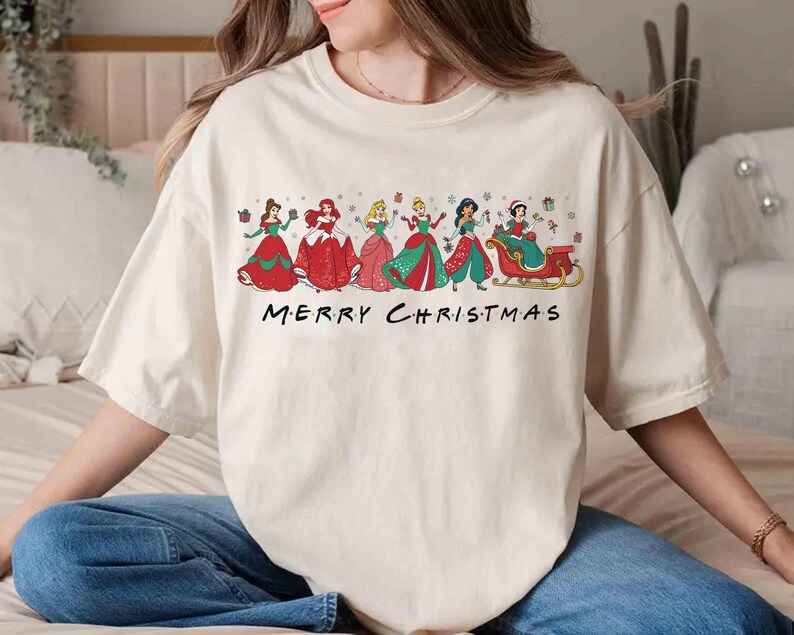 May include: Cream-colored t-shirt featuring a festive design with several princesses in holiday attire. The princesses are depicted in colorful dresses, with the text "MERRY CHRISTMAS" below them. A fun and festive holiday tee.
