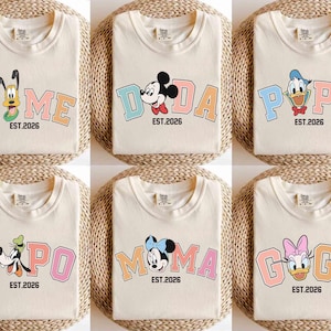 May include: Cream-colored t-shirts with colorful cartoon character designs. Each shirt features a different character and a word like "NOME", "DADA", "POPA", "POPO", "MOMA", and "GOGI", along with "EST.2026". The shirts are folded on a woven surface.