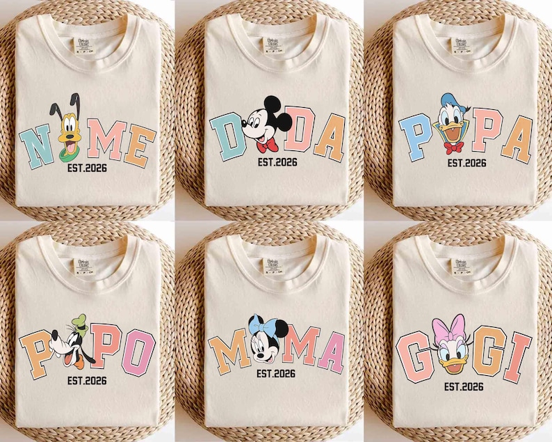 May include: Cream-colored t-shirts with colorful cartoon character designs. Each shirt features a different character and a word like "NOME", "DADA", "POPA", "POPO", "MOMA", and "GOGI", along with "EST.2026". The shirts are folded on a woven surface.