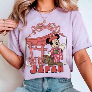 May include: A lilac-colored t-shirt featuring a graphic of Minnie Mouse in a kimono, holding a suitcase, and standing in front of a Japanese torii gate. The word "JAPAN" is printed in red block letters below the graphic.