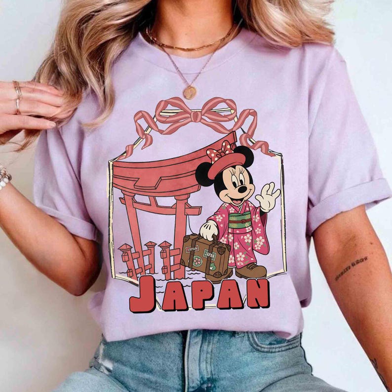 May include: A lilac-colored t-shirt featuring a graphic of Minnie Mouse in a kimono, holding a suitcase, and standing in front of a Japanese torii gate. The word "JAPAN" is printed in red block letters below the graphic.