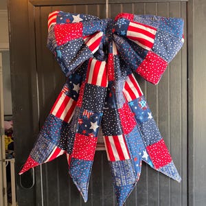 May include: A large, patriotic bow made of fabric patches. The bow features red, white, and blue patterns, including stars and stripes. The bow is hanging on a dark door.