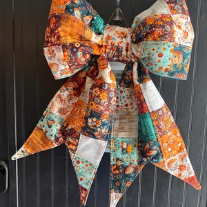 May include: A large decorative bow made from quilted fabric featuring a patchwork pattern of autumn-themed prints including pumpkins, fall leaves, sunflowers, and butterflies in shades of orange, teal, brown, cream, and yellow with long tails.
