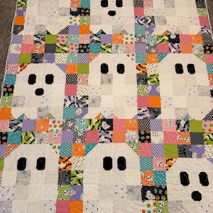 May include: A rectangular patchwork quilt featuring eight white ghost figures with black eyes and mouths. The background consists of a grid of small squares in various colours and patterns, including orange, purple, green, and black with skull motifs.