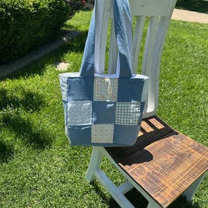 May include: A denim patchwork tote bag with blue and patterned squares. The bag has denim handles and a white lining. It is hanging on a white wooden chair, set on a grassy lawn.