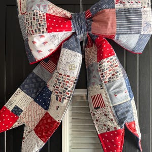 May include: A large, patriotic bow made of patchwork fabric, featuring red, white, and blue patterns. The bow has a quilted appearance with various designs, including stars, flags, and houses. It is likely a decorative item for a door or wall.