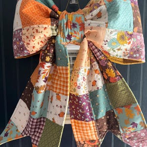 May include: A large decorative bow made from quilted fabric featuring a patchwork design. The fabric displays various autumn-themed patterns including orange pumpkins, colorful fall leaves, sunflowers, and plaid prints in shades of orange, brown, green, and cream.