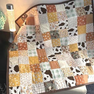 May include: A patchwork quilt with a brown border, featuring squares of various patterns including cow print, floral, and gingham in shades of brown, yellow, and cream. The quilt is draped over a chair, showcasing its design and texture.