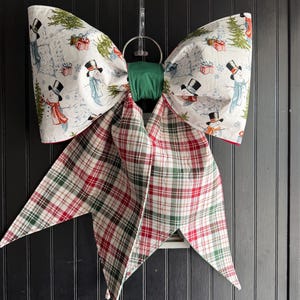 May include: A large fabric bow featuring a white background with a repeating pattern of snowmen wearing top hats and scarves, interspersed with green pine trees and red gift boxes. The bow has long, pointed tails in a red, green, and white plaid.