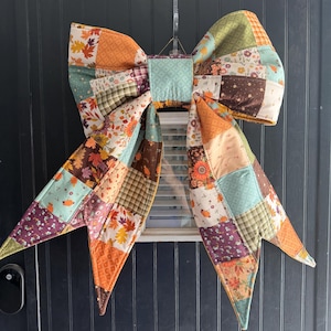 May include: A large decorative bow made from quilted fabric featuring a patchwork design. The fabric squares display various patterns including autumn leaves, floral prints, and geometric textures in shades of orange, brown, green, and cream.