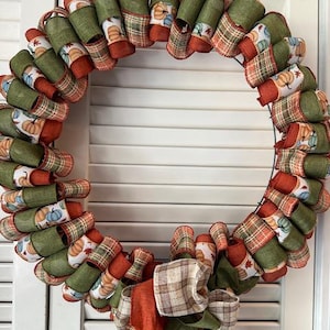 May include: A fall-themed wreath with a circular design. The wreath features green, orange, and white ribbons. Some ribbons display pumpkin patterns, while others have plaid designs. A large bow at the bottom adds a decorative touch.