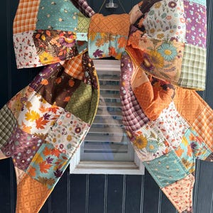 May include: A large decorative bow constructed from quilted fabric patches featuring a variety of autumn-themed patterns, including orange and brown maple leaves, sunflowers, plaid, and floral prints in shades of teal, cream, and mustard yellow.
