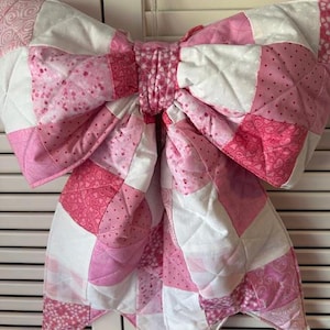 May include: A large, quilted bow in shades of pink and white. The bow is made of fabric squares with various patterns, including polka dots and hearts. The bow is likely a decorative item, possibly for a wall or door.