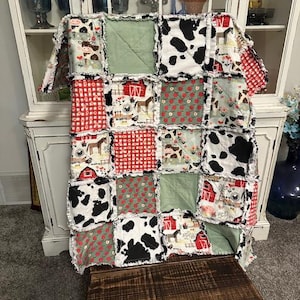 May include: A patchwork quilt with a farm animal theme. The quilt features squares of fabric with cow spots, red gingham, green, and farm animal prints. The quilt is displayed on a wooden bench in front of a white cabinet.