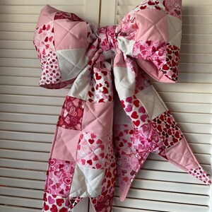 May include: A large, quilted bow in shades of pink, white, and red, featuring a patchwork design with heart patterns. The bow is made of fabric and has a diamond-stitched texture. It is a decorative item.