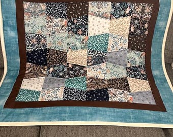 May include: A patchwork quilt with a teal border and a brown inner border. The quilt's center features a variety of patterned squares in shades of teal, brown, navy, and cream. The quilt is displayed on a gray couch.