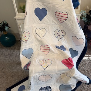 May include: A white quilt with a grid pattern, featuring various heart designs in blue, pink, and patterned fabrics. The quilt has a dark blue border and is draped over a surface.