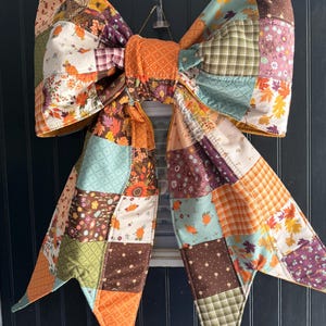 May include: A large decorative bow constructed from a patchwork of quilted fabric squares. The fabric features various autumn-themed patterns including orange pumpkins, fall leaves, brown plaid, and floral prints in shades of orange, green, and brown.