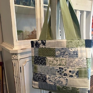 May include: A patchwork tote bag with a green handle. The bag is made of various fabric patterns in shades of blue, green, and white. The bag is hanging in front of a white cabinet.
