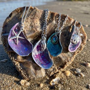 May include: Four seashell necklaces with silver chains and pendants. The pendants are made from iridescent shells in shades of purple and blue, with starfish, sand dollar, and other beach-themed charms. The necklaces are displayed on a rock on a sandy beach.