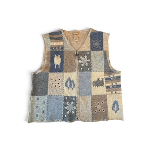 May include: A sleeveless zip-up vest featuring a patchwork design in shades of blue, beige, and grey. The knit garment displays various embroidered motifs including snowflakes, trees, animals, and geometric patterns with visible stitching.