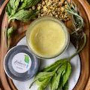 Peut inclure: Un plateau en bois présente un petit pot en verre de baume jaune, une petite boîte avec une étiquette et des herbes fraîches. Des brins de menthe, de camomille et de romarin entourent le pot et la boîte sur une assiette blanche.