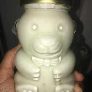 Op de afbeelding: Een heldere glazen pot in de vorm van een teddybeer, gevuld met een lichtgekleurde substantie. De pot heeft een goudkleurige deksel en een vlinderdasdetail. De beer-vormige pot wordt in een hand gehouden.