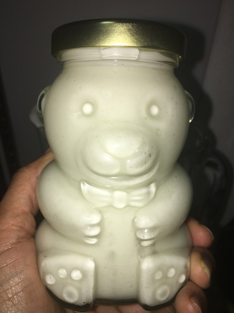 Op de afbeelding: Een heldere glazen pot in de vorm van een teddybeer, gevuld met een lichtgekleurde substantie. De pot heeft een goudkleurige deksel en een vlinderdasdetail. De beer-vormige pot wordt in een hand gehouden.