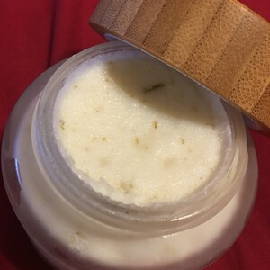 Op de afbeelding: Een heldere glazen pot gevuld met een romige, gebroken witte substantie, mogelijk een cosmetisch product. De pot heeft een houten deksel dat gedeeltelijk open is en de inhoud onthult. De achtergrond is een effen rode stof.