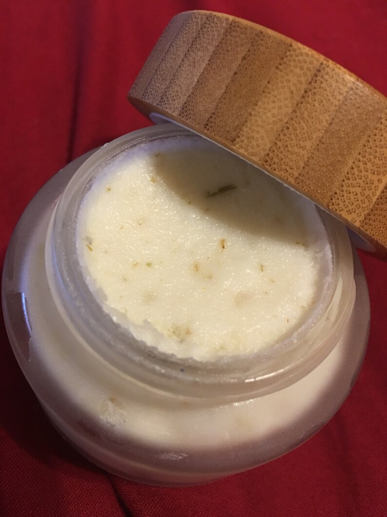 Op de afbeelding: Een heldere glazen pot gevuld met een romige, gebroken witte substantie, mogelijk een cosmetisch product. De pot heeft een houten deksel dat gedeeltelijk open is en de inhoud onthult. De achtergrond is een effen rode stof.
