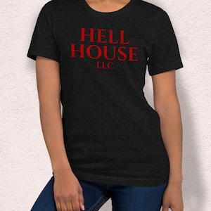 May include: Dark gray t-shirt with the red text "HELL HOUSE LLC". The shirt has short sleeves and a crew neck. The text is centered on the chest.