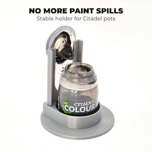 Puede incluir: Un soporte gris para botes de pintura con el texto "NO MORE PAINT SPILLS" y "Stable holder for Citadel pots". El soporte tiene una base redonda y una cuna para sujetar un bote de pintura Citadel Colour.