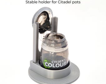 May include: A gray paint pot holder with the text "NO MORE PAINT SPILLS" and "Stable holder for Citadel pots". The holder has a round base and a cradle to hold a Citadel Colour paint pot.